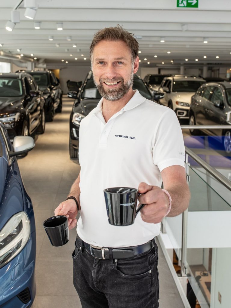 Mann i hvit t-skjorte foran mange biler hos en bilforhandler