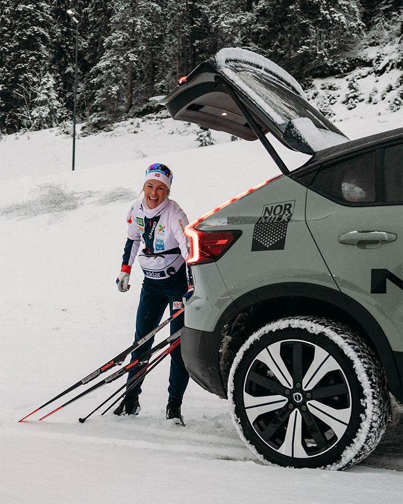 Kvinne står ved bagasjeluken til en grønn suv med skiutstyr