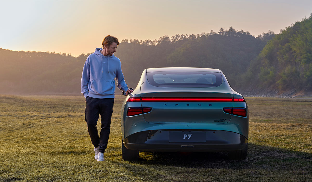 Grønn Xpeng P7 på med baksiden mot kamera på grønn slette i solnedgang. Mann står på siden av bilen med arm på en dør.